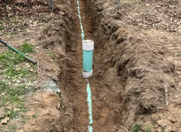 A long, narrow trench dug in a residential yard between a chain-link fence and a sidewalk, showing a teal pipe running along the bottom and a short