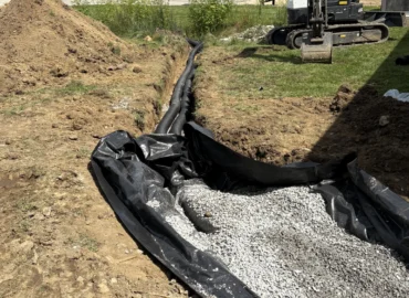 A trench dug in the dirt, lined with black plastic fabric and filled with small gray drainage gravel